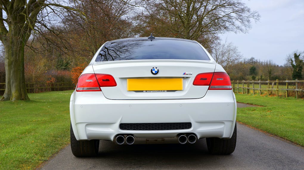 Elegant white BMW M3 parked on a scenic countryside road in the UK, emphasizing rear design and lush surroundings.