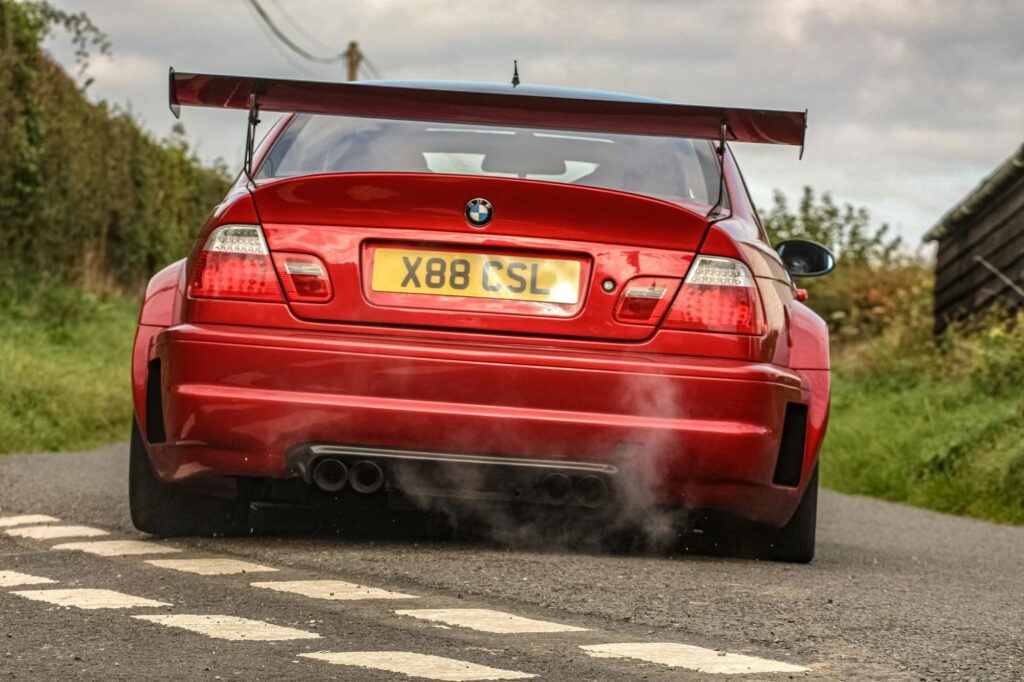 Why the BMW E46 M3 Is a Terrible First Enthusiast Car 2 Red BMW E46 M3 sportscar captured on a country road, showcasing wide rear arches and massive spoiler