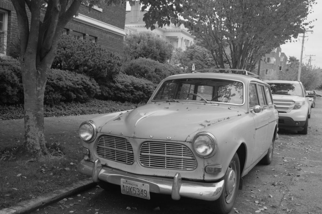 Classic Volvo Amazon parked on a leaf-covered street in a suburban neighborhood.